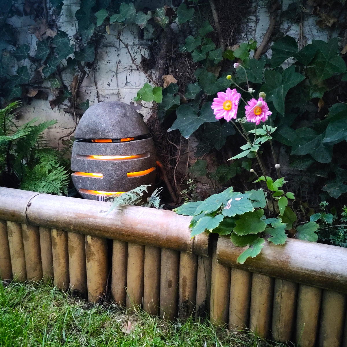 Laterne aus Naturstein mit warm beleuchteten Schlitzöffnungen neben Gartenblumen